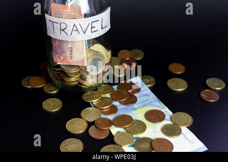 Transparent en verre pot étiqueté voyage manuscrite. Billets et pièces à l'intérieur et l'extérieur du pot. Fond noir isolé. Banque D'Images