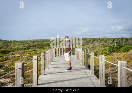 Homme barbu portant une casquette vue depuis l'avant, regardant vers le bas, dans une distance debout sur une passerelle en bois au milieu de la nature. Banque D'Images