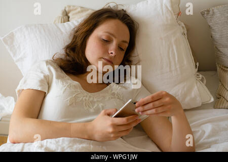 Jeune femme au lit le matin et la lecture des messages sur smartphone Banque D'Images