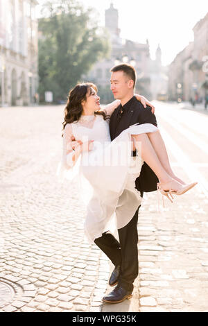 Belle asiatique jeunes mariés sur le mariage marche à travers les rues de la vieille ville européenne. Man holding woman sur les mains. Banque D'Images