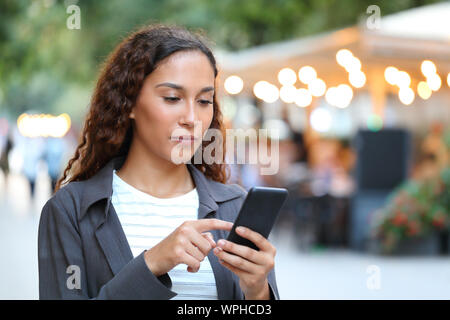 Serious mixed race woman using smart phone marche dans la rue avec les lumières de la ville dans l'arrière-plan Banque D'Images