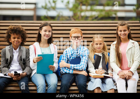 Portrait de l'école accueillant les enfants avec des livres assis sur le banc et smiling at camera outdoors Banque D'Images