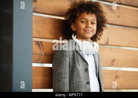 Portrait d'écolier africain avec des cheveux bouclés portant veste et un casque à la caméra à tout en s'appuyant sur le mur extérieur en bois Banque D'Images