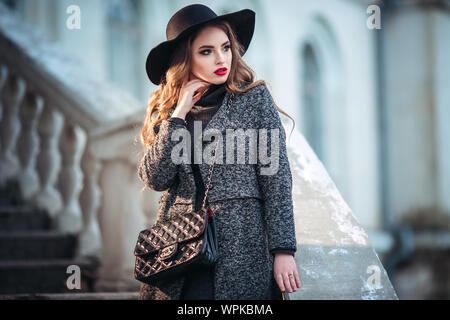 Belle jeune fille avec perfect make-up, rouge lèvres, portant un chapeau noir et gris manteau, robe noire, posant dans evning city. Banque D'Images