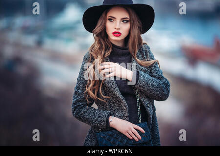 Belle jeune fille avec perfect make-up, rouge lèvres, portant un chapeau noir et gris manteau, robe noire, posant dans evning city. Banque D'Images