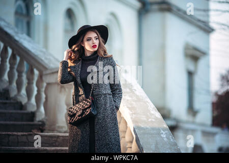 Belle jeune fille avec perfect make-up, rouge lèvres, portant un chapeau noir et gris manteau, robe noire, posant dans evning city. Banque D'Images