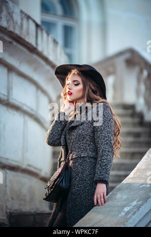 Belle jeune fille avec perfect make-up, rouge lèvres, portant un chapeau noir et gris manteau, robe noire, posant dans evning city. Banque D'Images