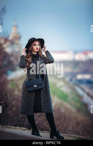 Belle jeune fille avec perfect make-up, rouge lèvres, portant un chapeau noir et gris manteau, robe noire, posant dans evning city. Banque D'Images