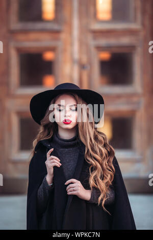 Belle jeune fille avec perfect make-up, rouge lèvres, portant un chapeau noir et gris manteau, robe noire, posant dans evning city. Banque D'Images