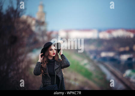 Belle jeune fille avec perfect make-up, rouge lèvres, portant un chapeau noir et gris manteau, robe noire, posant dans evning city. Banque D'Images