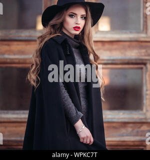 Belle jeune fille avec perfect make-up, rouge lèvres, portant un chapeau noir et gris manteau, robe noire, posant dans evning city. Banque D'Images