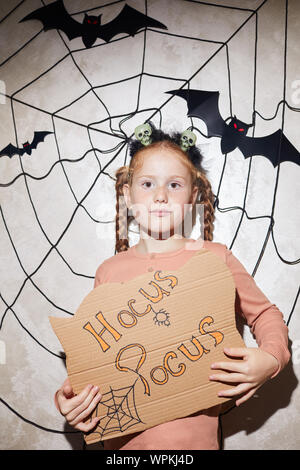 Portrait of cute girl holding Hocus Pocus placard et posant à huis clos avec d'araignée et les chauves-souris dans l'arrière-plan Banque D'Images