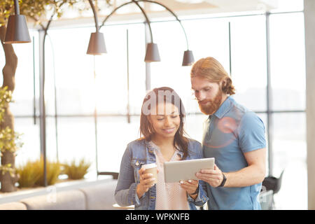 Jeune homme barbu regardant quelque chose sur tablet pc avec femme souriante avec un verre de café tout en se tenant à l'restaurant Banque D'Images