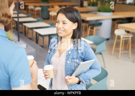 Young woman in casual clothing holding tablet pc de boire du café et de parler au jeune homme tandis qu'ils standing in cafe Banque D'Images