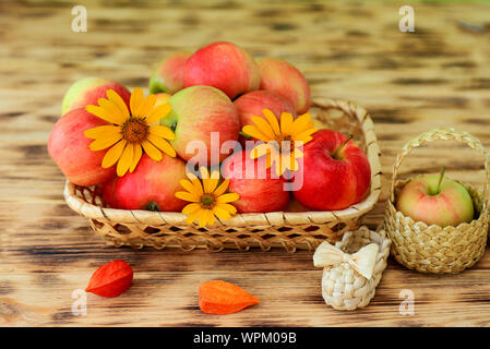 Le rouge des pommes mûres et de fleurs d'automne se trouvent dans une assiette sur une table en bois. Wicker bast chaussures et des pommes dans un panier. L'alimentation saine et le mode de vie Banque D'Images