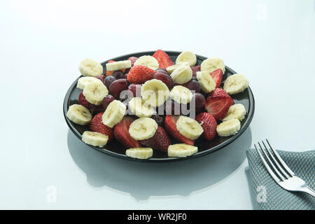 Banane Raisins et strawbery tranches sur une plaque noire sur une surface brillante et blanche avec une fourche sur le côté et l'arrière-plan blanc. Banque D'Images