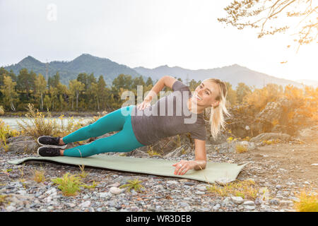 Belle fille blonde de sourire et de faire un échauffement allongé sur le sol en pierre dans la pose de la planche latérale, appuyé sur le coude à l'extérieur dans le mountain vues Banque D'Images