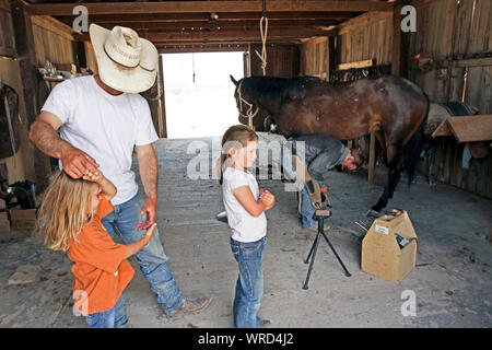 Jouant avec un éleveur de sa fille dans une grange sur un ranch dans l'ouest du Texas, alors qu'un cowboy travaille sur un fer à cheval. Banque D'Images