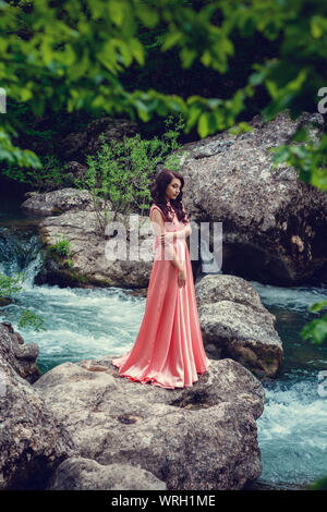 Sorcière de la rivière, assis sur un rocher dans la rivière. Robe rose, une image à la mode fabuleux.tonifiant. Couleur créative. Belle danseuse dans une forêt mystique b Banque D'Images