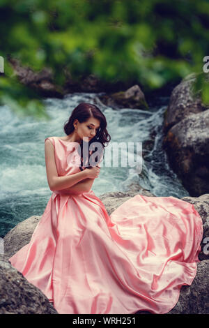 Sorcière de la rivière, assis sur un rocher dans la rivière. Robe rose, une image à la mode fabuleux.tonifiant. Couleur créative. Belle danseuse dans une forêt mystique b Banque D'Images