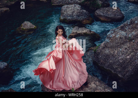 Sorcière de la rivière, assis sur un rocher dans la rivière. Robe rose, une image à la mode fabuleux.tonifiant. Couleur créative. Belle danseuse dans une forêt mystique b Banque D'Images