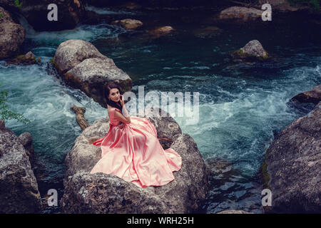 Sorcière de la rivière, assis sur un rocher dans la rivière. Robe rose, une image à la mode fabuleux.tonifiant. Couleur créative. Belle danseuse dans une forêt mystique b Banque D'Images