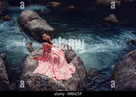 Sorcière de la rivière, assis sur un rocher dans la rivière. Robe rose, une image à la mode fabuleux.tonifiant. Couleur créative. Belle danseuse dans une forêt mystique b Banque D'Images