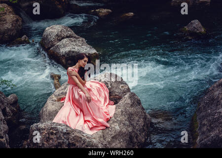 Sorcière de la rivière, assis sur un rocher dans la rivière. Robe rose, une image à la mode fabuleux.tonifiant. Couleur créative. Belle danseuse dans une forêt mystique b Banque D'Images