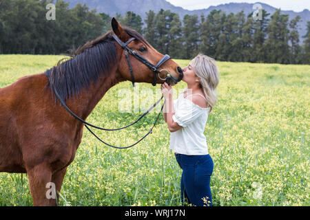 Woman kissing son cheval arabe alezan sur son nez, debout en face de l'autre, en plein air avec champ de fleurs jaunes. Banque D'Images