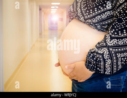 Selective focus on femme anonyme pregnant belly à la naissance, des problèmes de mise au point clinique de couloir de l'hôpital sur l'arrière-plan, des contrôles médicaux réguliers concept. Banque D'Images