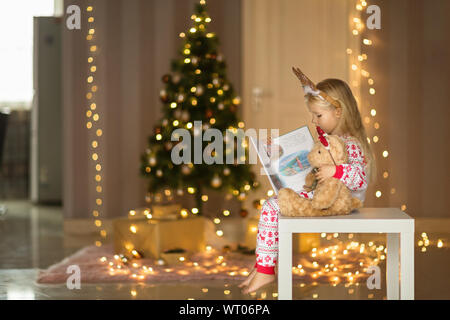 Jolie petite fille avec de longs cheveux blonds en pyjama de noël livre de lecture avec nounours. Décorations de Noël autour de. Heureux moments de la petite enfance Banque D'Images