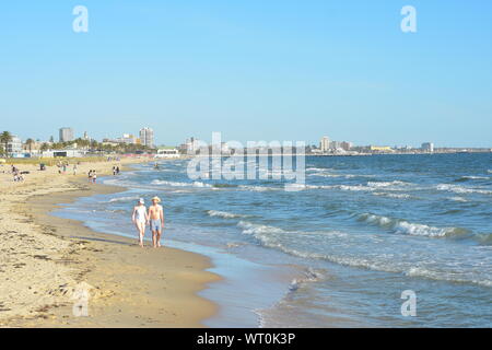 Les gens qui marchent le long de la plage de sable fin baignée par les vagues de surf océanique avec Melbourne cityscape in background. Banque D'Images