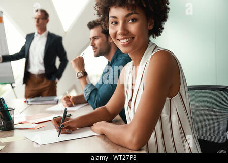 Cheerful businesswoman. Femme Jeune est looking at camera et souriant alors que son patron, debout près de tableau blanc et discuter de quelque chose avec l'équipe. Le travail d'équipe. Réunion Banque D'Images