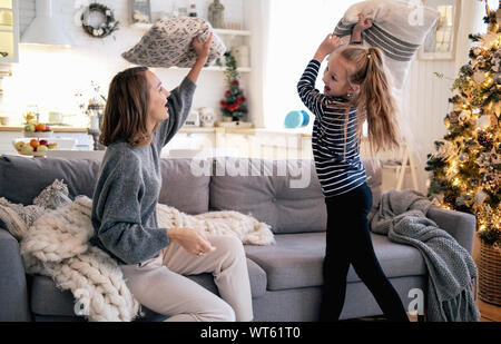 Mère et fille jouer pillow fight en canapé à la maison. Le matin de Noël, vacances, nouvelle année, des couleurs douces. Banque D'Images