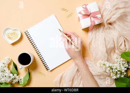 Haut de la vue, télévision laïques 24 avec des fleurs lilas, soie, tasse à café, boîte-cadeau, human hand holding golden pen et l'écriture dans le bloc-notes de papier vierge. Banque D'Images