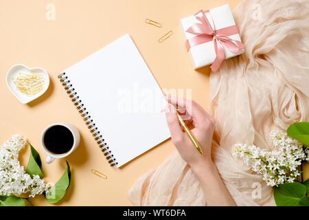 Haut de la vue, télévision laïques 24 avec des fleurs lilas, soie, tasse à café, boîte-cadeau, human hand holding golden pen et l'écriture dans le bloc-notes de papier vierge. Banque D'Images