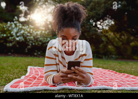 Smiling Relaxed young african american woman lying on blanket sur pelouse dans le parc à l'aide de téléphone mobile - étudiant sur leur mobile à College Park Banque D'Images