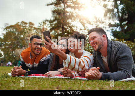 Smiling multi ethnic group of friends lying on Green grass smiling tout en tenant sur smartphone selfies dans le parc - très nature vert Banque D'Images