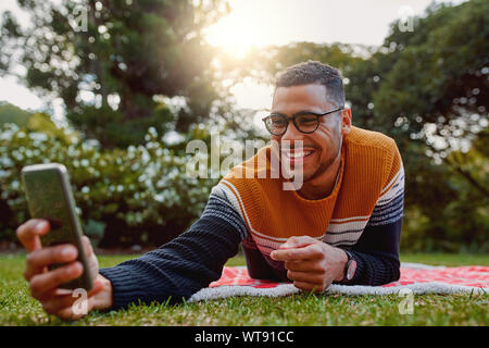 Portrait of a smiling young african american man lying on blanket sur l'herbe verte dans le parc en tenant sur selfies téléphone mobile au collégial - universitaire Banque D'Images