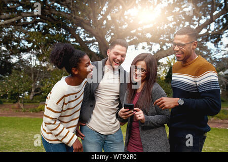 Groupe diversifié d'happy friends Standing together de rire et à la recherche à smart phone dans le parc - très branché habillé Banque D'Images