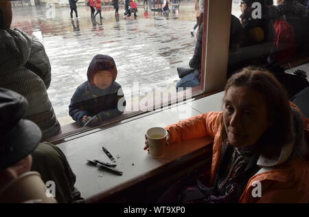 Enfant chinois à regarder les touristes prenant le thé à travers une fenêtre d'une cour extérieure froide et pluvieuse à Beijing, Chine Banque D'Images