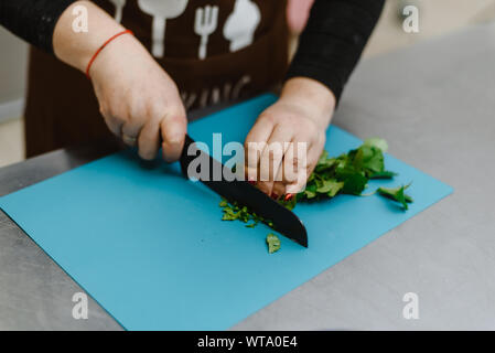 La jeune fille coupe verts, oignons, persil et divers assaisonnements avec un couteau sur une planche à découper. Femme cuire salade pour la cuisson. Banque D'Images