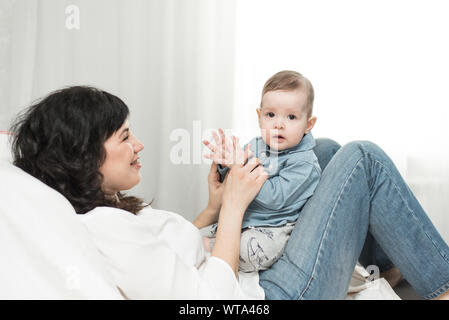 Jeux amusants avec un an mère tout-petit.heureux famille positif à la maison, jeune mère aux cheveux noirs se trouve sur le canapé et s'amuse avec sa fille pl Banque D'Images