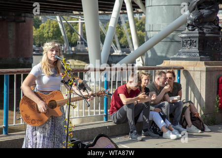 Chanteur musicien ambulant avec la guitare sur la rive sud de Londres avec une famille de touristes assis contre une balustrade de la Tamise avoir quelque chose à manger. Banque D'Images