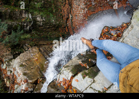 Les jambes de l'homme en jeans et bottes marron assise sur le bord à la cascade à Banque D'Images