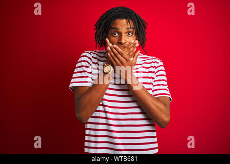 Jeune homme avec des dreadlocks afro portant des T-shirt à rayures sur fond rouge isolé permanent choqué couvrant la bouche avec les mains pour l'erreur. Con Secret Banque D'Images