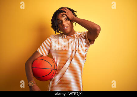 Sportif avec des dreadlocks Afro holding basketball ball sur fond jaune isolé a souligné avec la main sur la tête, choqué par la honte et la surprise fac Banque D'Images