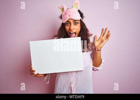 Jeune femme portant un masque de sommeil et de pyjama holding banner sur fond rose isolé très heureux et excité, lauréat expression célébrant la victoire s Banque D'Images