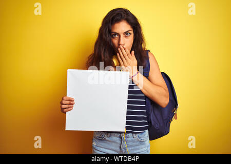 Belle jeune femme étudiante holding banner debout sur fond jaune isolé couvrir la bouche à part choqué avec honte pour l'erreur, d'expression Banque D'Images