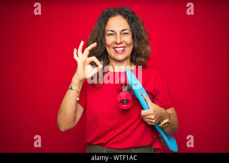 L'âge moyen du patineur senior woman holding skateboard rouge sur fond isolé faisant signe ok avec les doigts, symbole excellent Banque D'Images